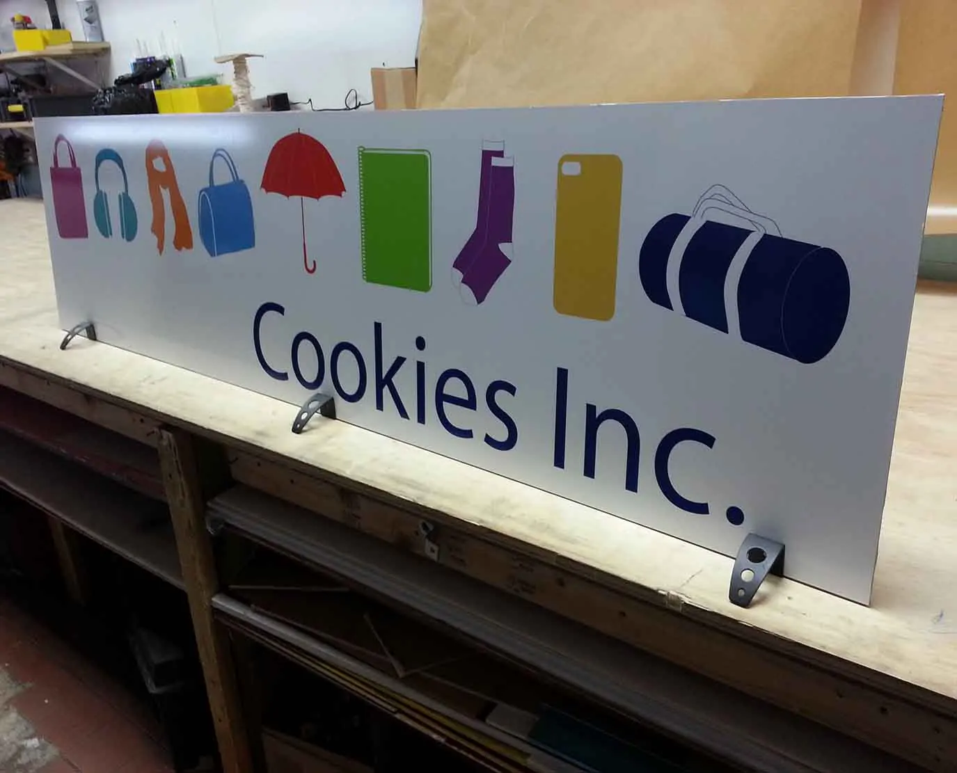 White aluminum sign panel for Cookies Inc. with colorful product icons on a shop bench before installation