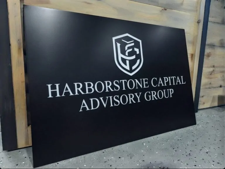 Large black Dibond sign panel with a brushed silver crest reading Harborstone Capital Advisory Group, propped up before installation