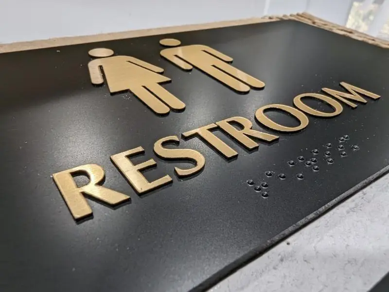 Black ADA restroom sign with gold raised letters and braille