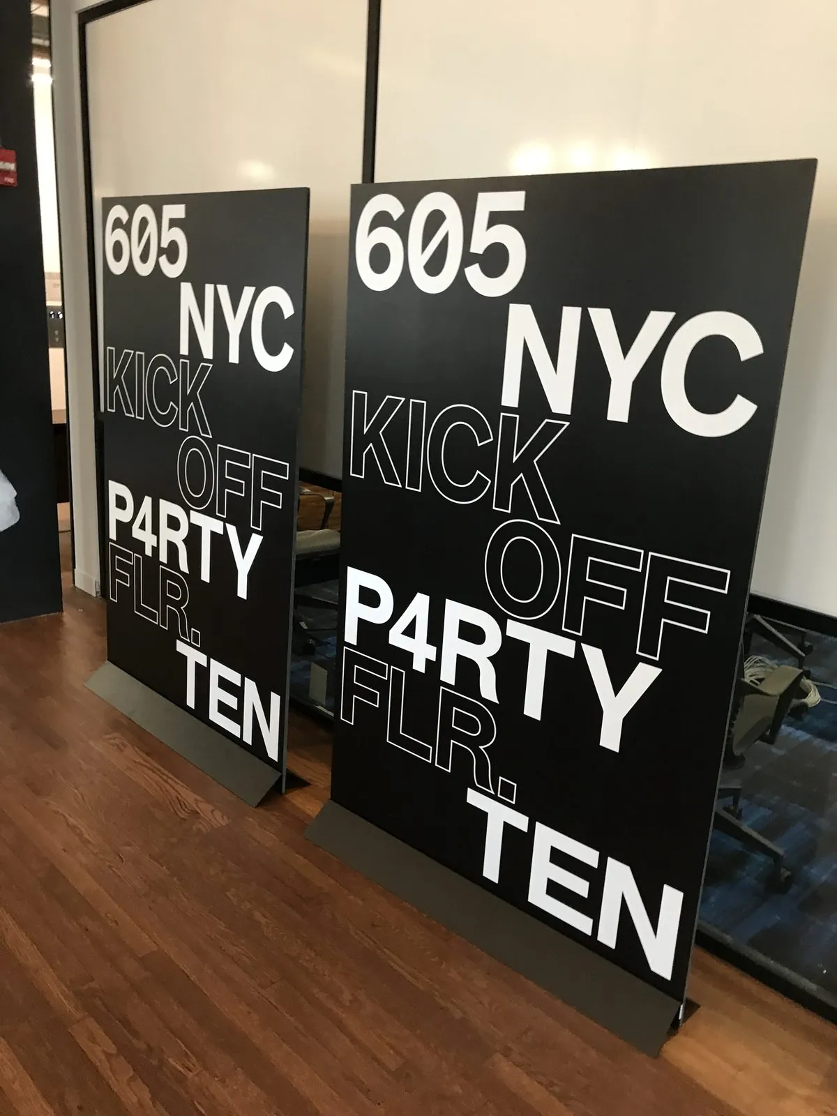 Two black freestanding event panels with large white type, standing on a wood floor