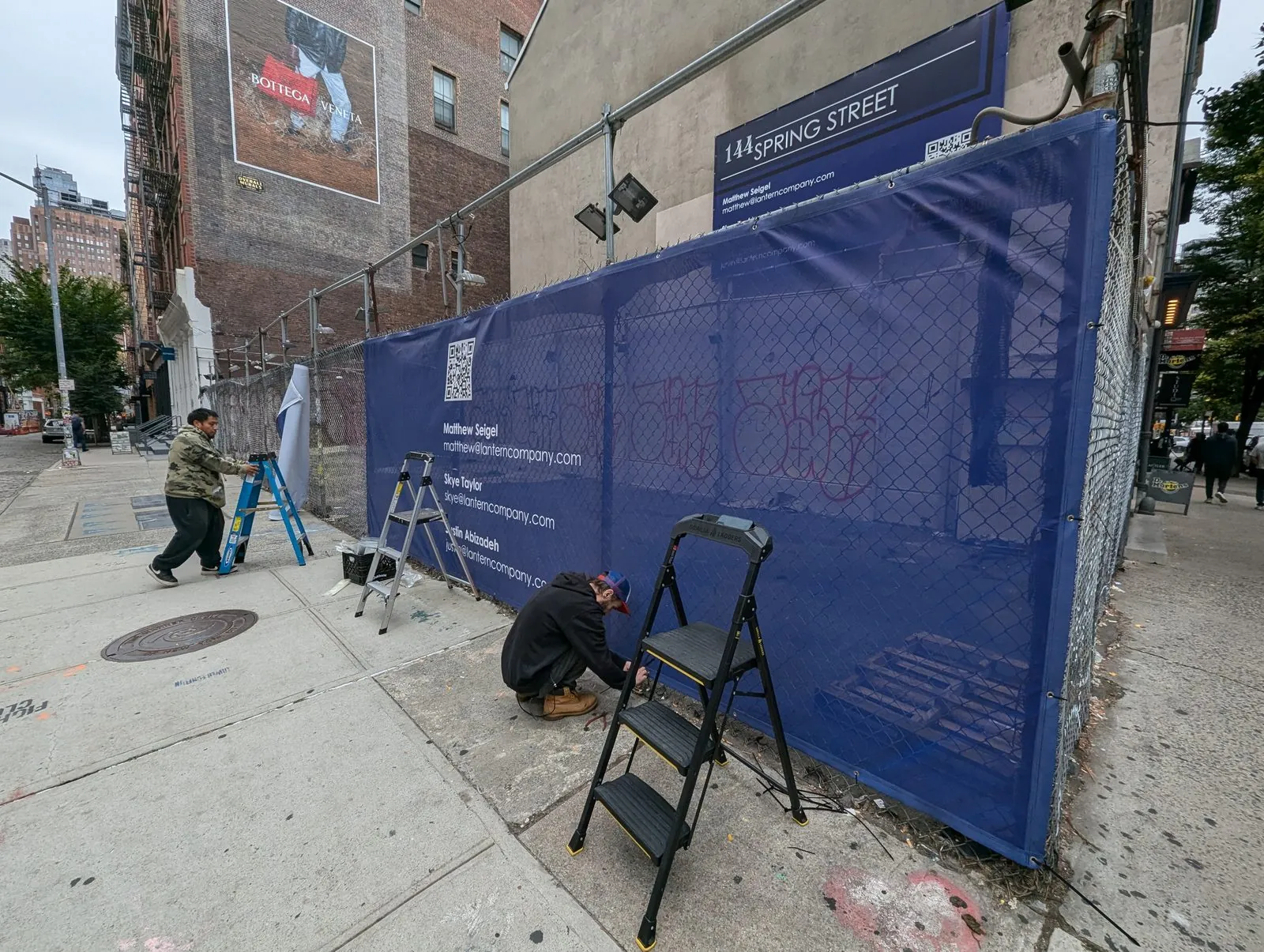 Crew tying a mesh banner to a chain link fence on a city sidewalk