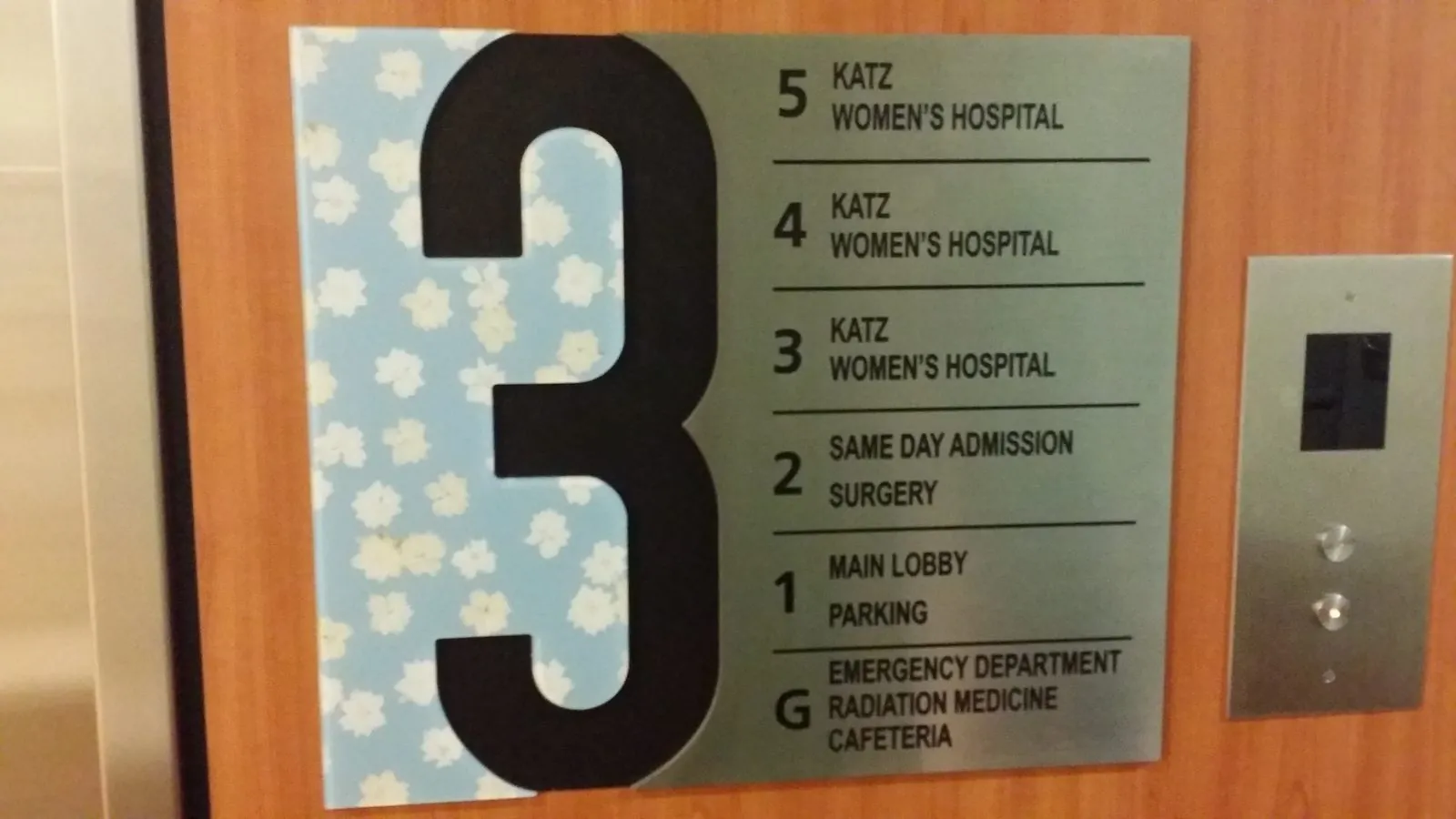 Elevator lobby sign with a tall black number 3 on a patterned panel and brushed metal floor listings beside the call buttons