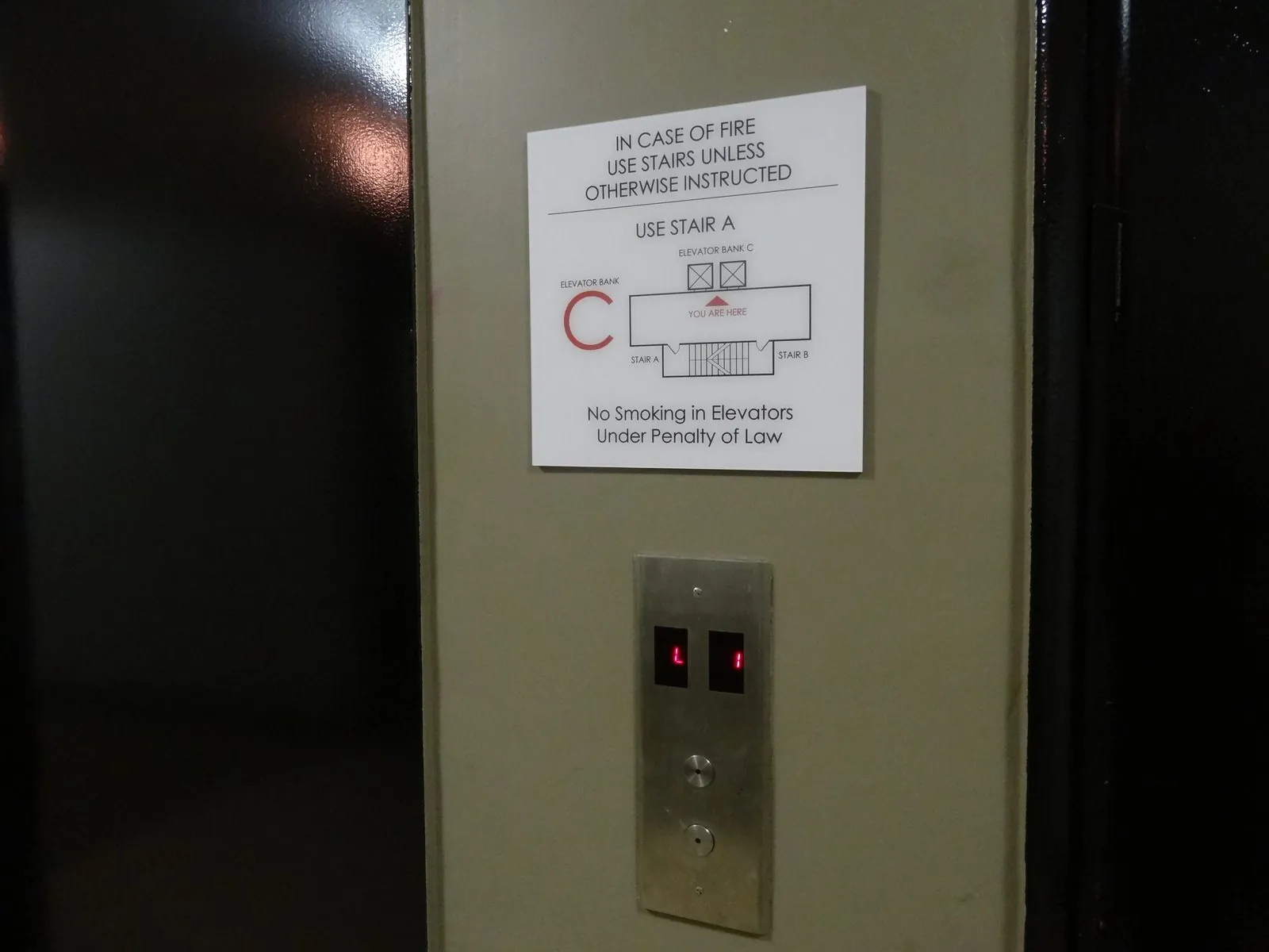 White elevator lobby sign with a red bank letter C and a small floor plan mounted above the elevator call buttons