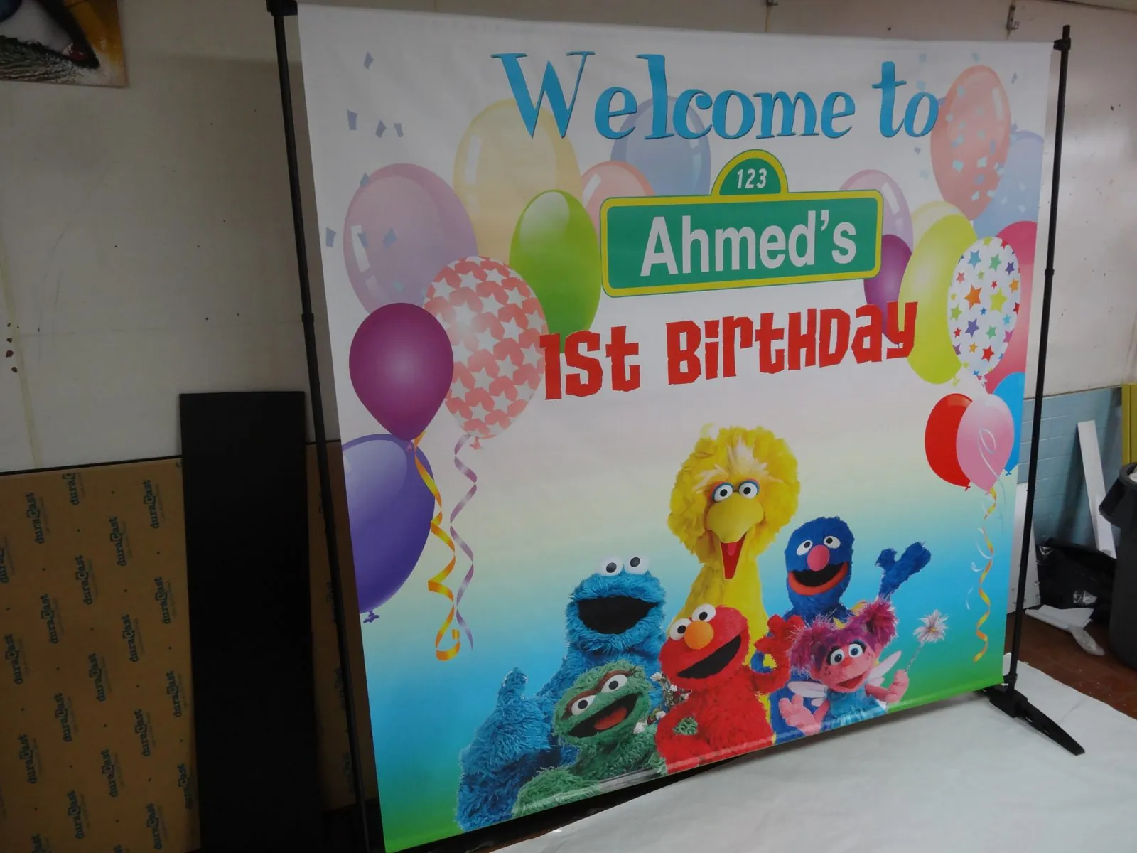 Colorful first birthday backdrop with balloons, a name banner and cartoon characters on a frame