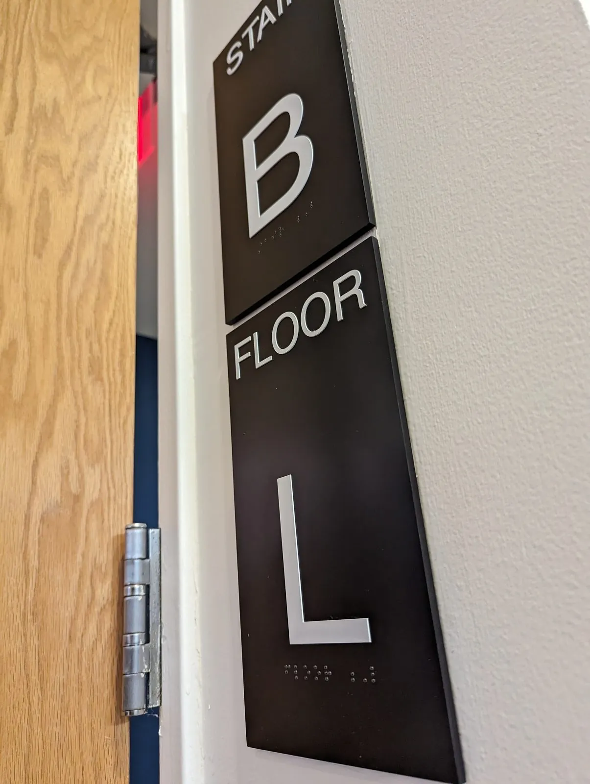Black Stair B and Floor L signs with raised white characters stacked beside a door frame