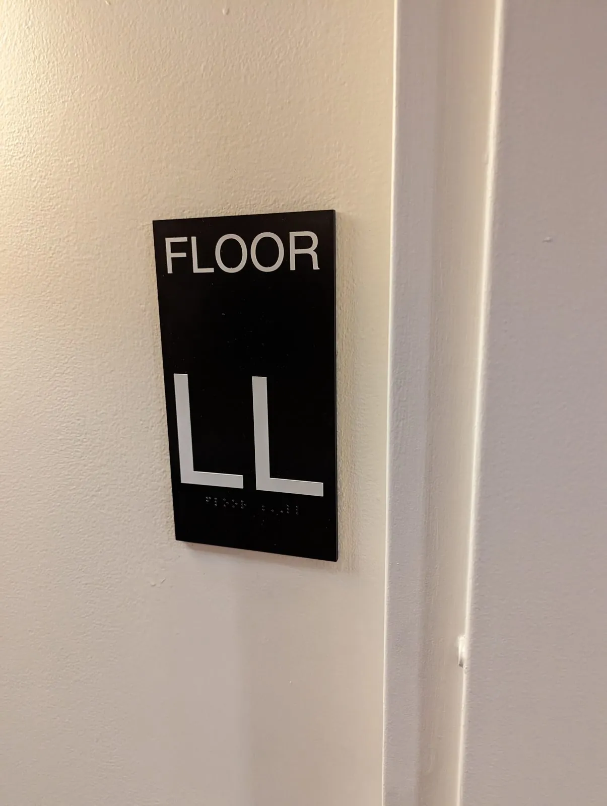 Black Floor LL sign with raised white letters and braille on a white wall by a door frame
