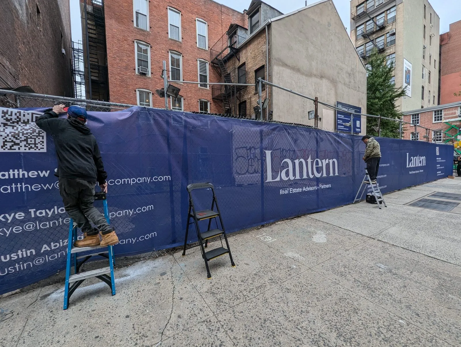 Installer on a step ladder tying a navy mesh banner to a construction fence