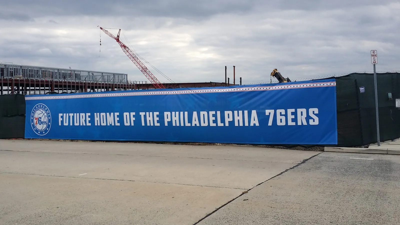 Long blue mesh banner on a construction fence announcing the future home of a sports team, with cranes behind