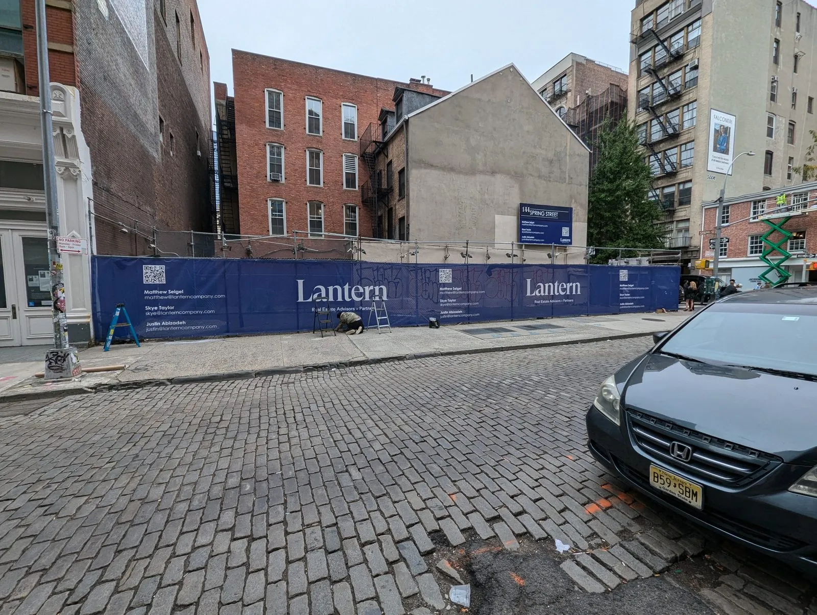 Long navy mesh banner printed with a logo and contact lines along a construction fence on a cobblestone street