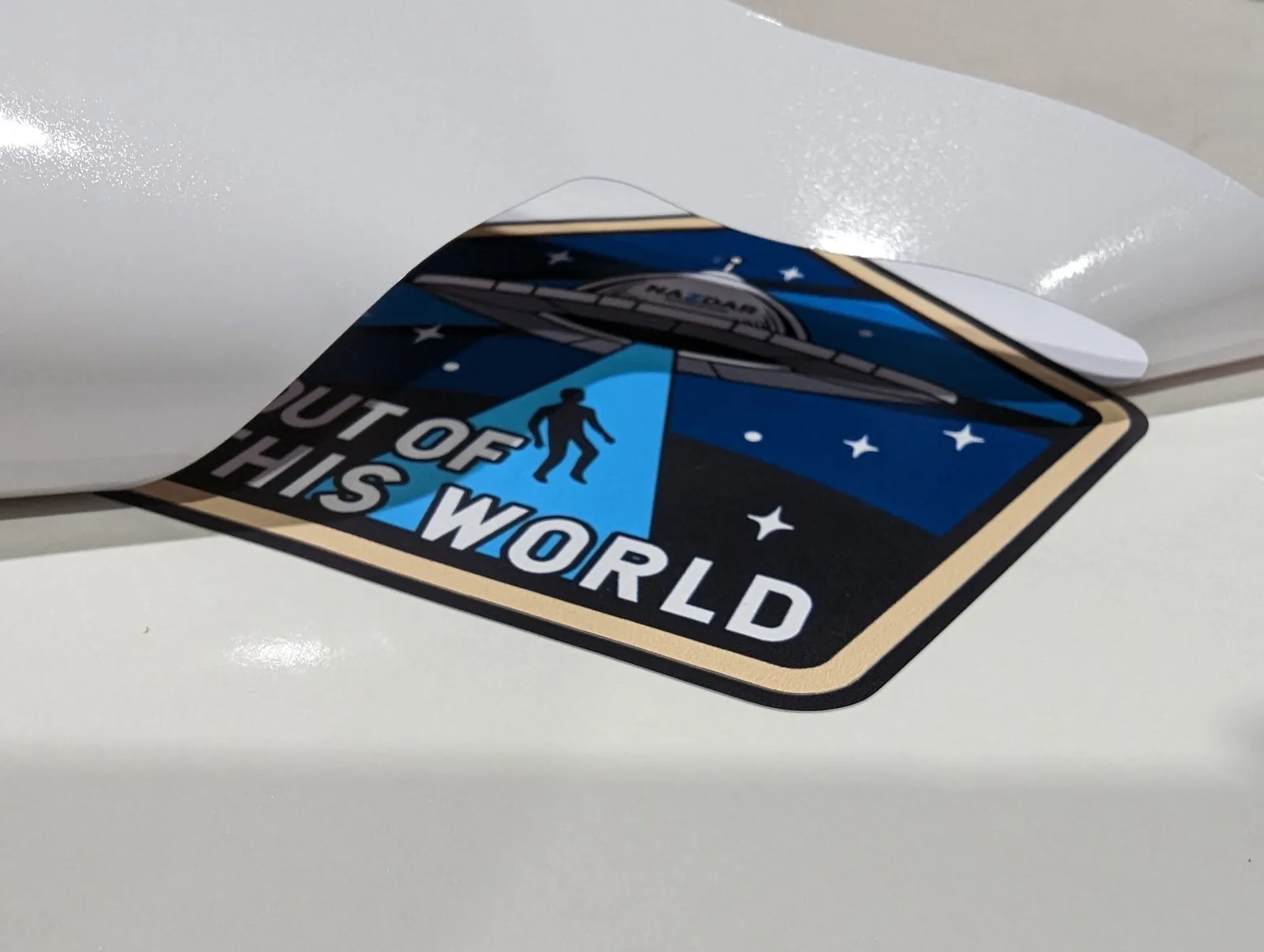 Full color badge decal reading Out of This World half peeled from its white backing liner