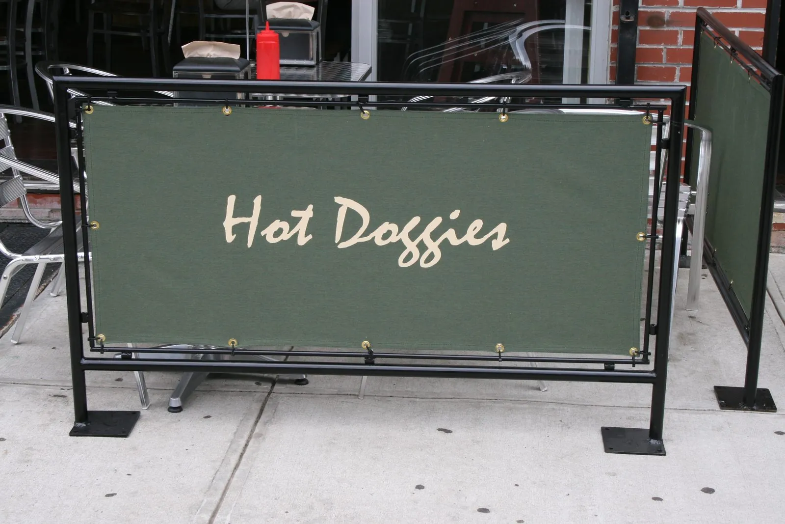 Green printed banner reading Hot Doggies laced into a black steel cafe barrier on a sidewalk