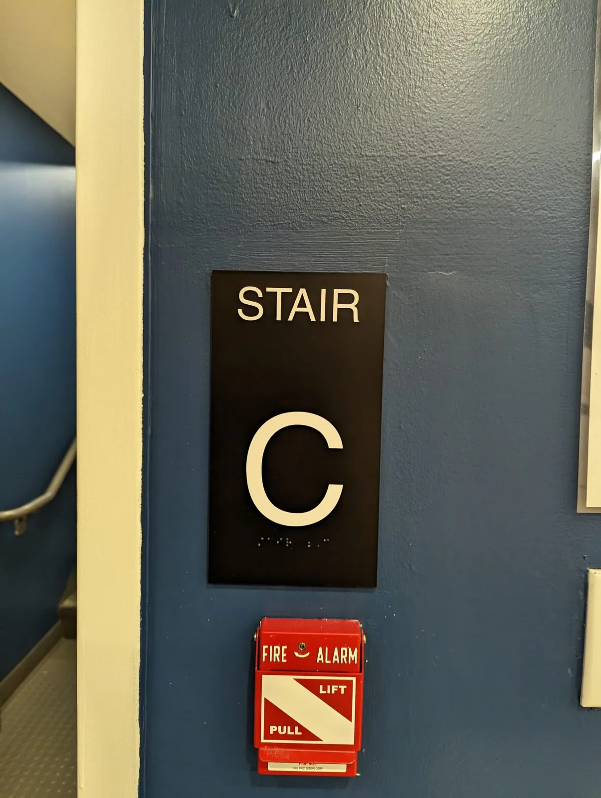 Black Stair C sign with braille above a red fire alarm pull station on a blue wall