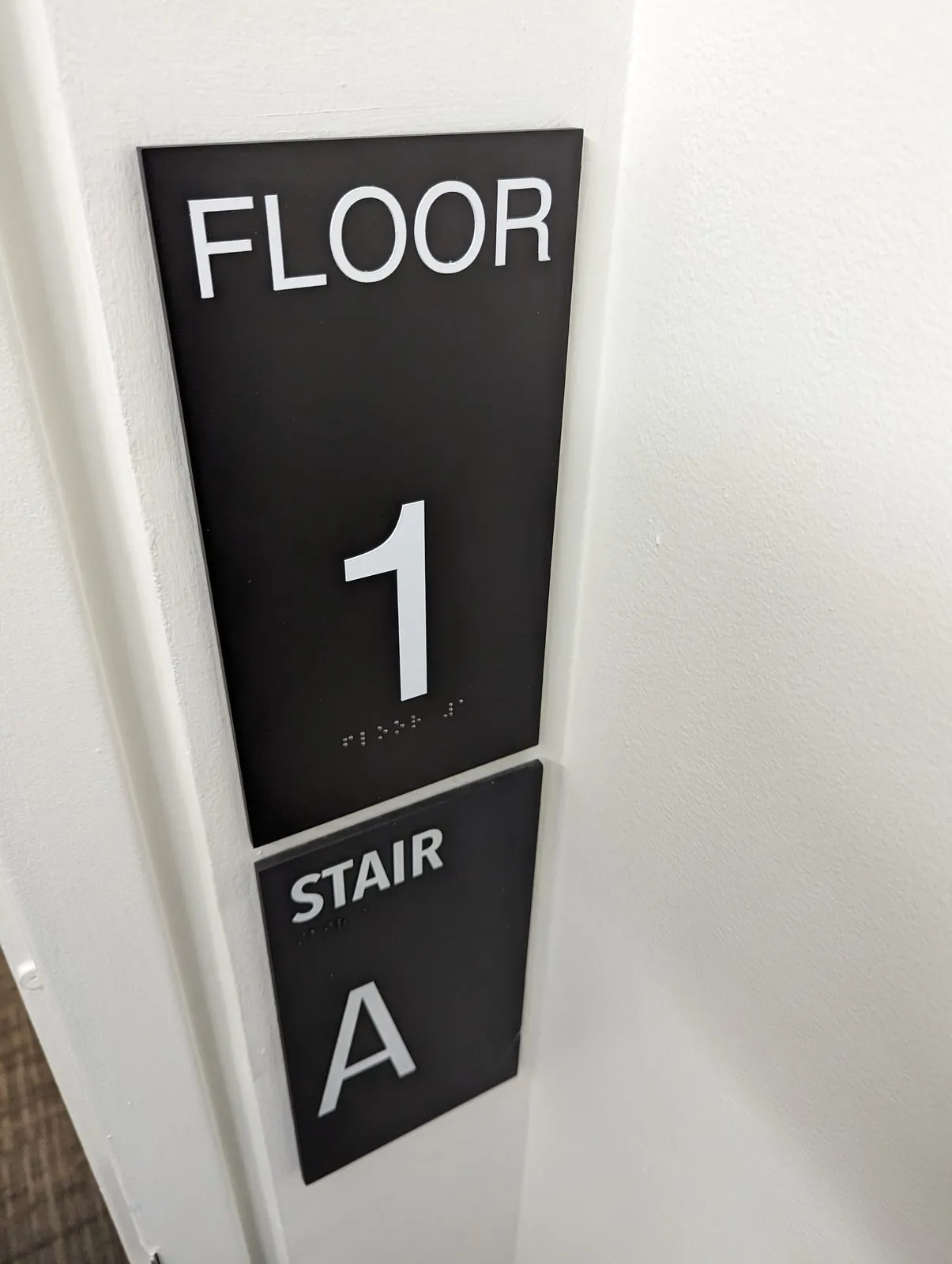 Black Floor 1 and Stair A signs with raised white characters and braille beside a door frame