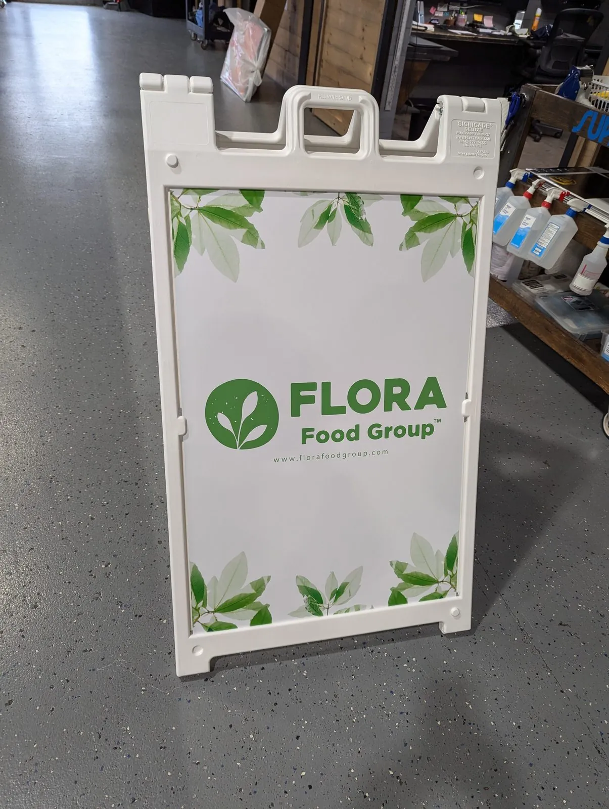 White plastic sandwich board sign with a green leaf border and a food company logo