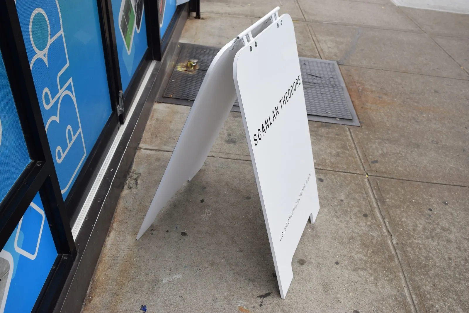 White plastic sandwich board seen from the side on a sidewalk beside a shop window