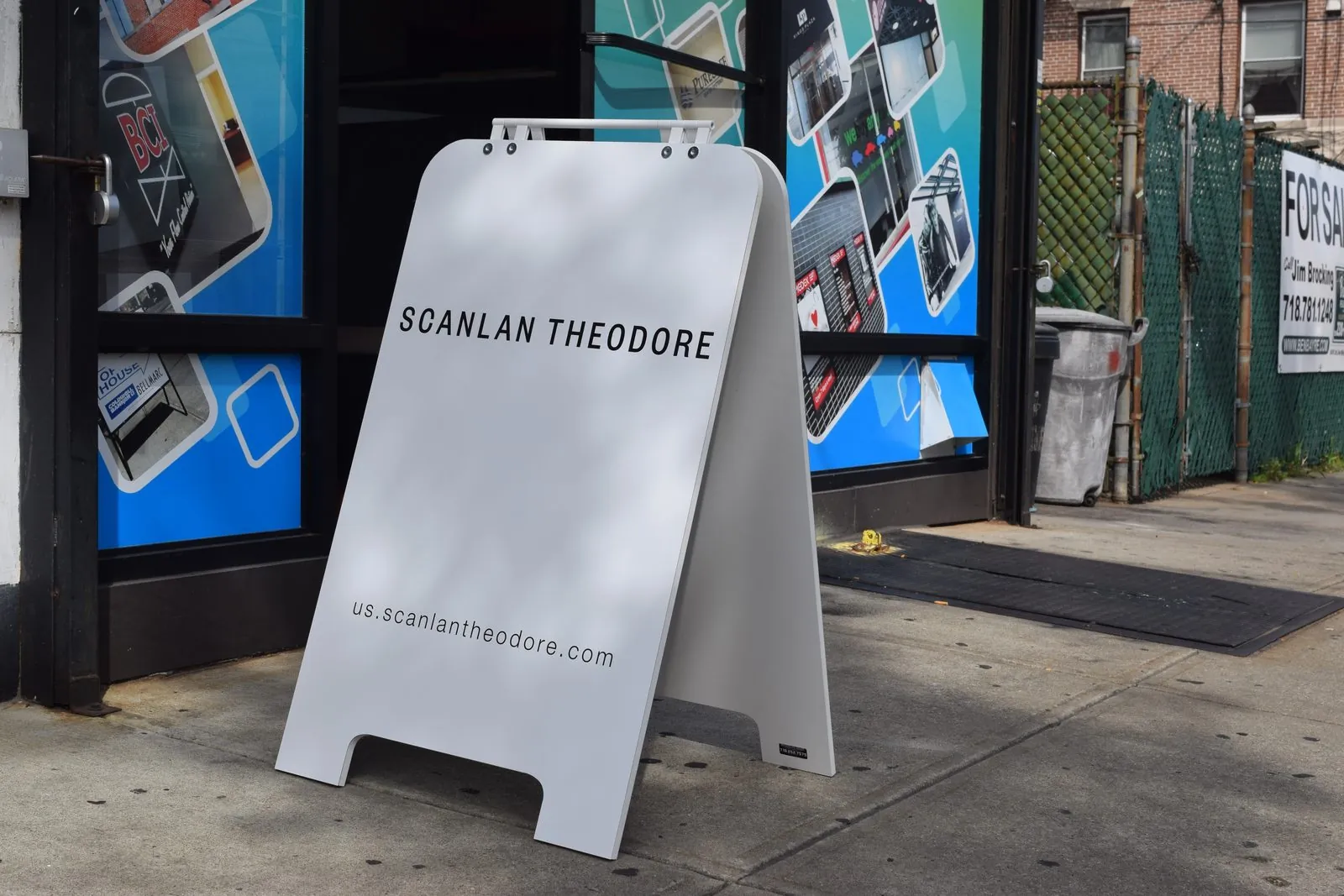 White plastic sandwich board with a black name on a sidewalk beside a storefront